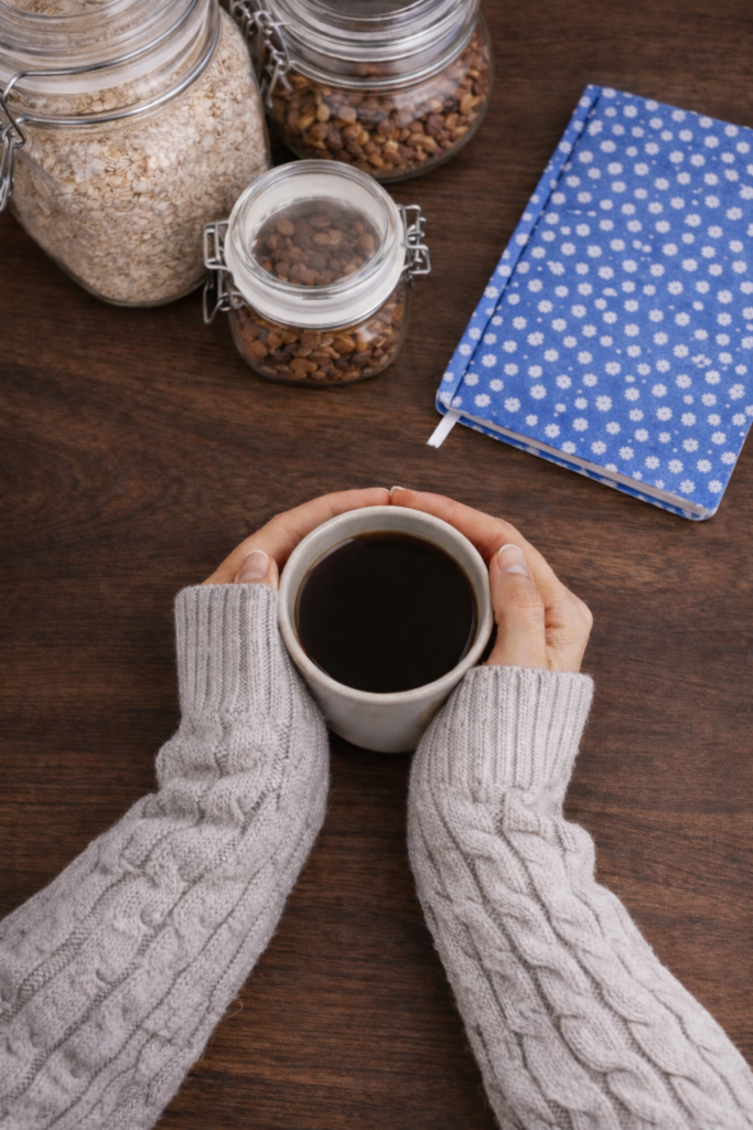 Hände mit Kaffeetasse in den Händen und Notizbuch und Glas mit Nüssen im Hintergrund